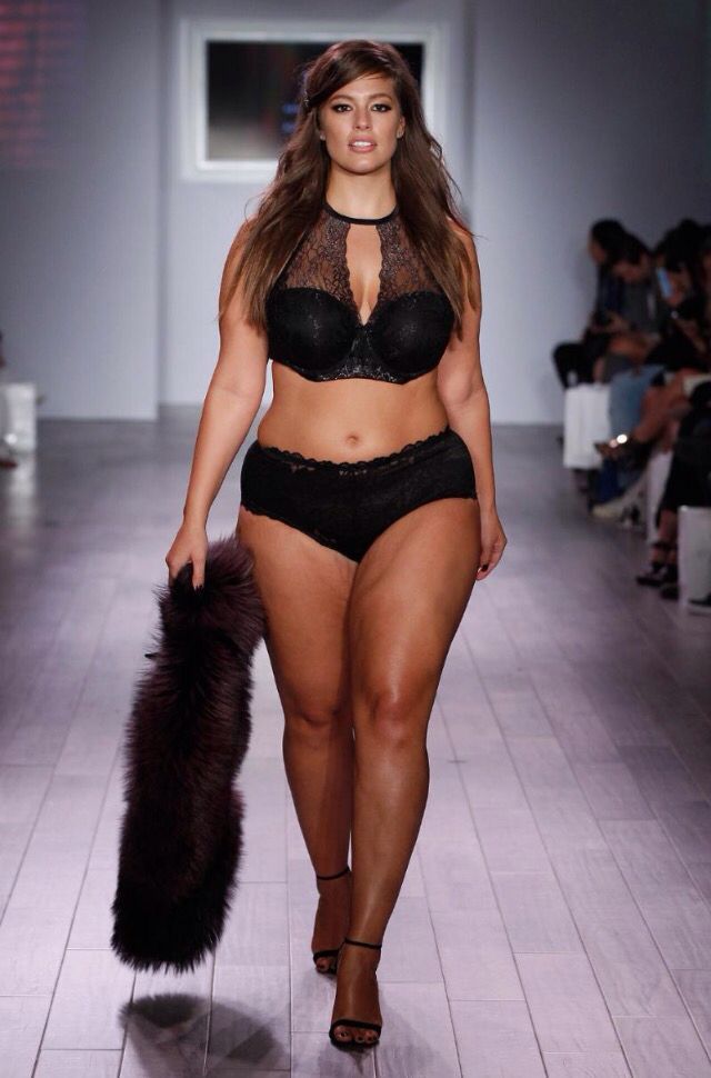 A model walking down the runway in black lingerie and ankle strap heels, holding a faux fur wrap.